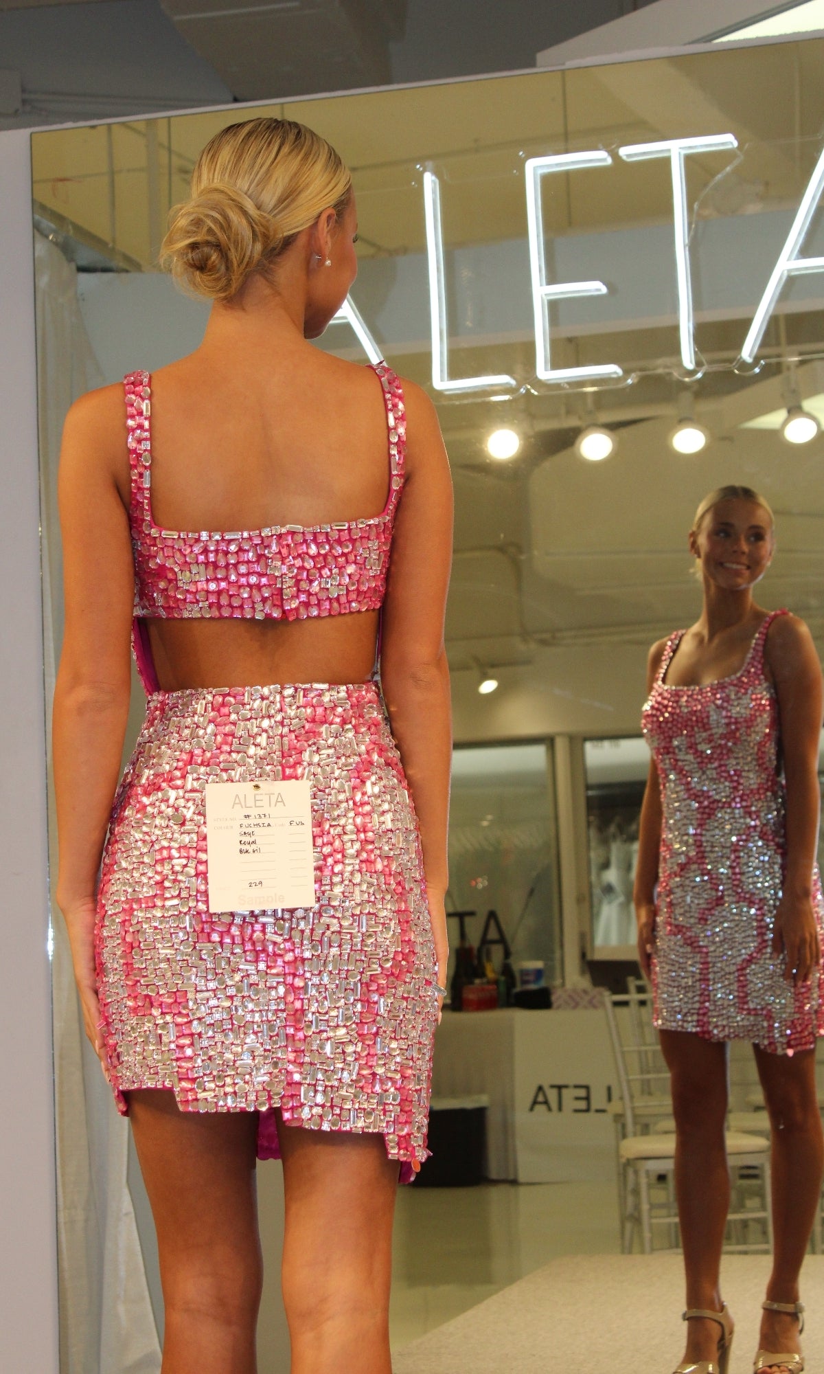 A blonde woman with a bun models the Short Homecoming Dress 1371 by Aleta, featuring pink and silver beading, an open back, and a tag, standing before a mirror showing the “ALETA” brand name.